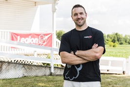 A young male veteran with short brown hair stands in front of a Legion branch in the summertime. He is wearing a black t-shirt with the Legion logo on it and jeans. His arms are crossed and he is smiling for the camera.